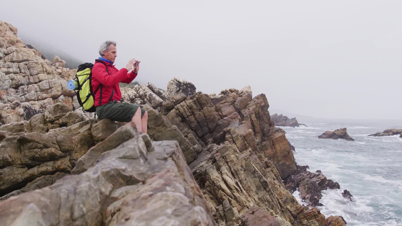 hombre excursionista sénior con mochila sentado en las rocas y tomando fotos en el teléfono inteligente mientras caminaba