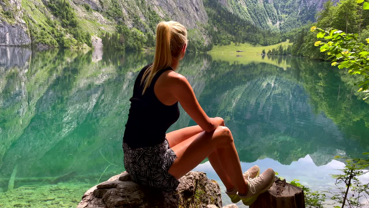 Attractive young woman enjoying crystal clear lake in nature with mountains in background - 4K Slow motion shot of Caucasian blond girl sitting on shore and enjoying wilderness