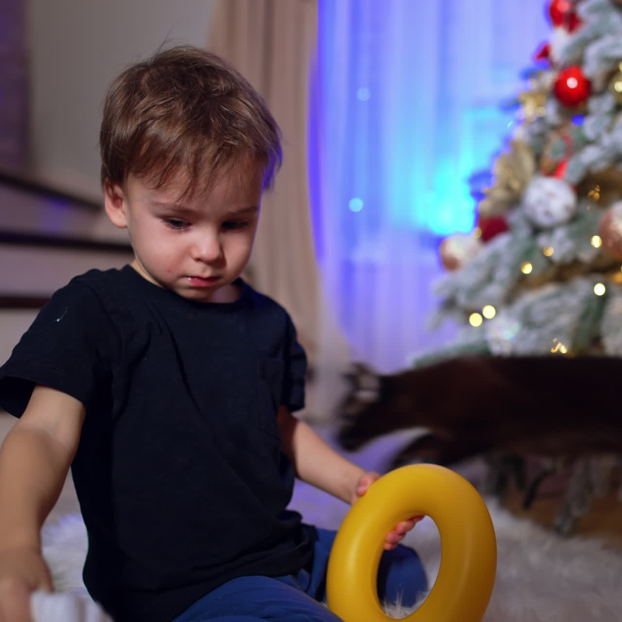 Two year old toddler takes off Santa cap. Funny baby puts yellow toy ring to his face. Christmas tree at backdrop.