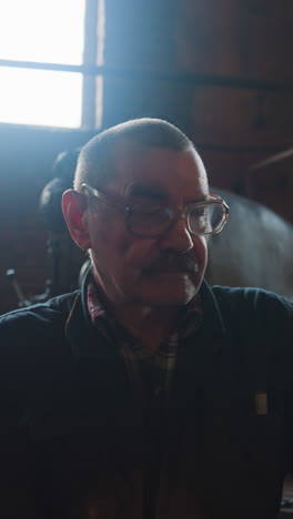 old labourer with spectacles sits by drilling machine in factory workshop. employee at workplace with vintage lathe equipment. metalworking technology