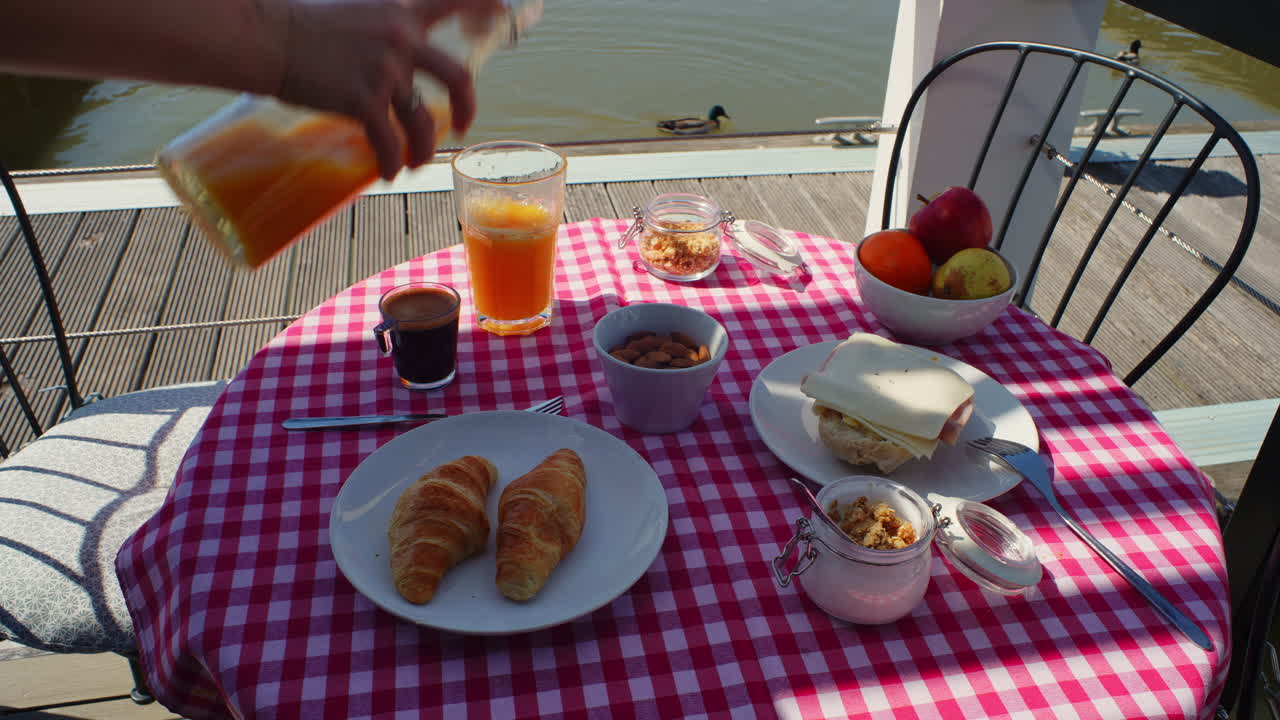Mano sirviendo jugo de naranja en una mesa de desayuno al aire libre junto al agua