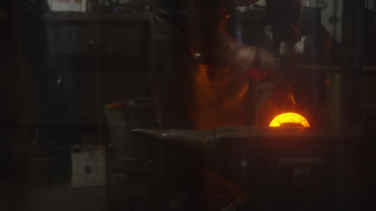 Blacksmith shaping a glowing chain link in a dark forge workshop with low light and dramatic atmosphere