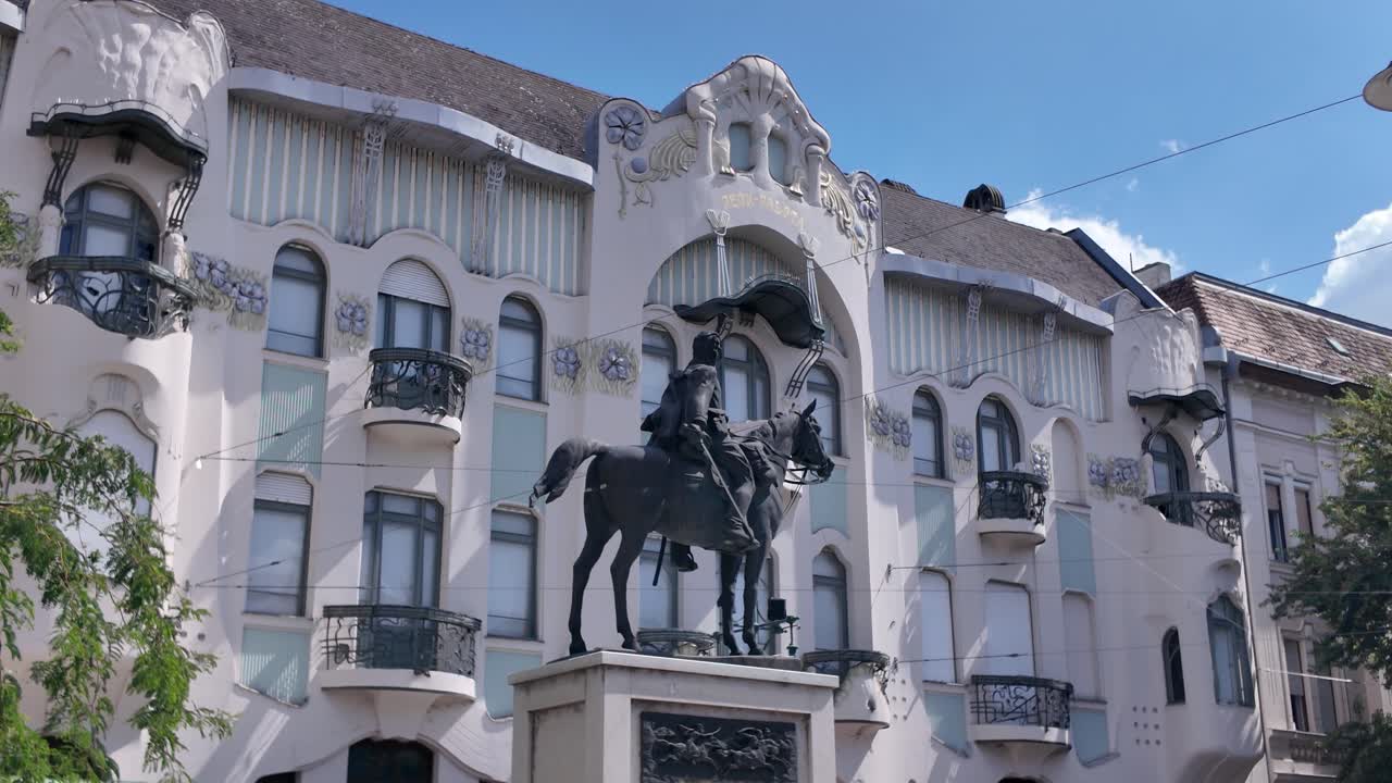 Pan across Reök Palace with a foreground monument in Szeged, capturing both heritage architecture and urban surroundings