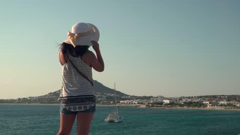 back of girl holding on to summer hat while looking out on ocean with boat and fishing town in distance