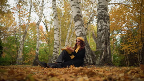 dama elegante en la soledad se apoya contra el árbol volviendo a través del libro pensativo, rodeada de hojas doradas de otoño, ella abre cuidadosamente la página, perdida en profunda contemplación