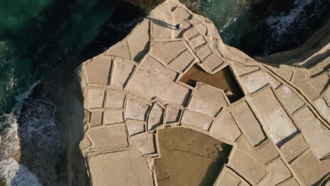 Aerial-high-angle-view-of-a-man-walking-across-pools-of-salt-pans-in-Xwejni-during-a-beautiful-sunset