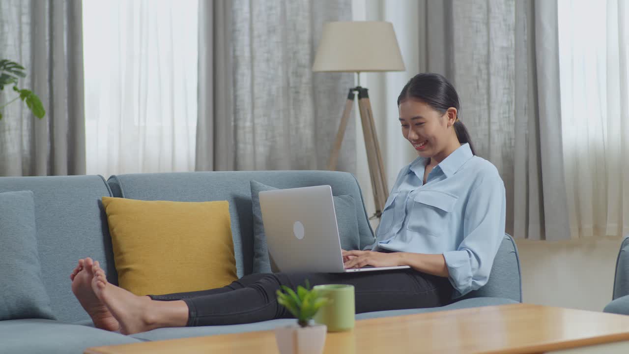 미소 짓는 아시아 여성의 몸이 가득 차 있으며, 거실의 소파에 누워있는 동안 노트북 컴퓨터에서 타이핑하고 있습니다.