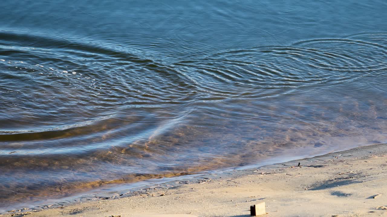 Small waves and ripples move across a calm river or lake, gently lapping against a sandy shoreline in bright natural daylight, captured in a steady wide shot
