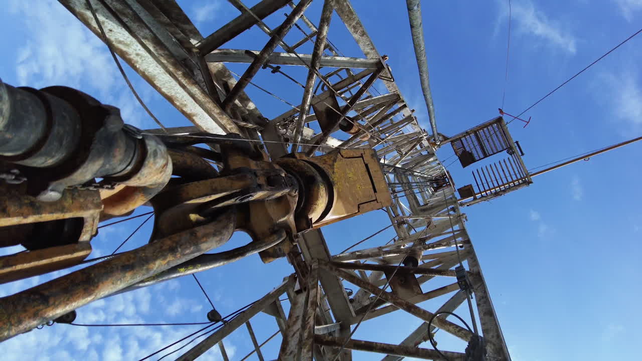 Footage at the base of a metal derrick for boring oil. Thick wires vibrate as the borer equipment works. Low angle view.