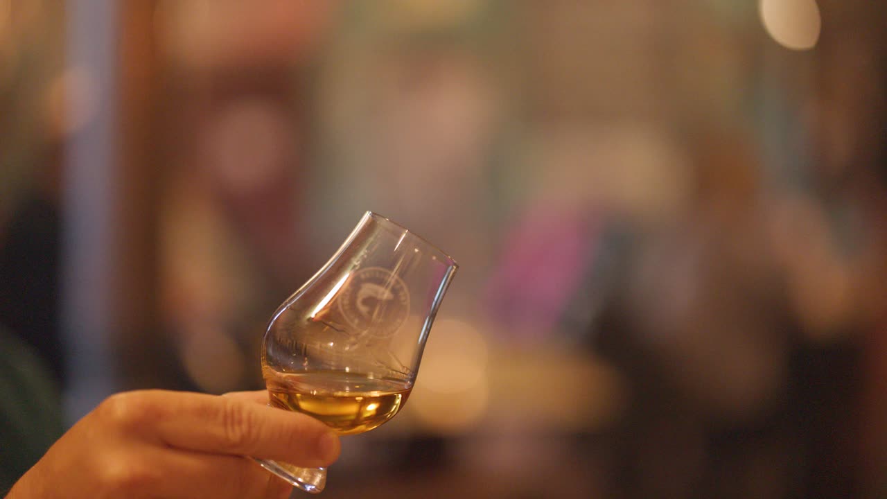 A hand gently swirls a whisky glass in a warmly lit pub, with a softly blurred background and subtle camera movement highlighting the drink