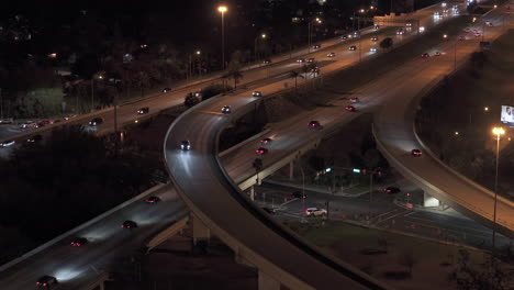 Nighttime aerial view of a curved multilane highway interchange with illuminated roads, palm trees, and flowing traffic under streetlights.