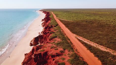 drone rastreando camino de tierra sobre acantilados rojos del lado del océano con playa de arena y agua azul