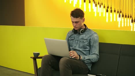 Attractive-Male-Sitting-On-Sofa-In-Well-Lit-Indoors-Space-Drinking-Coffee,-Working-On-His-Laptop