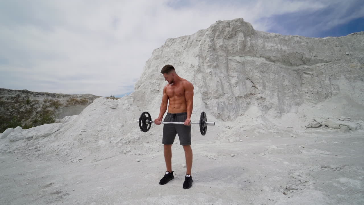 Young guy in shorts trains his muscular body outdoors. Bodybuilder lifting metal barbell in white mountains. Slow motion.