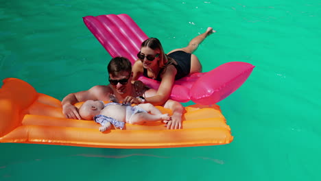 swimming in the pool with an infant. mother lies on the pink mattress and baby is on the orange one held by dad.