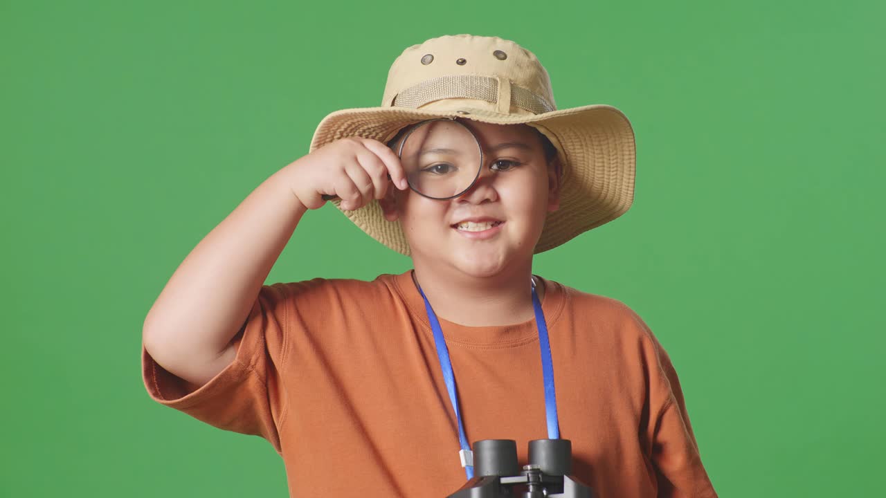 모자와 안경을 쓴 아시아 관광 소년은 창을 통해 녹색 화면 배경에서 무언가를 조사합니다. 소년 연구자, 여행 관광 모험 개념, 근접
