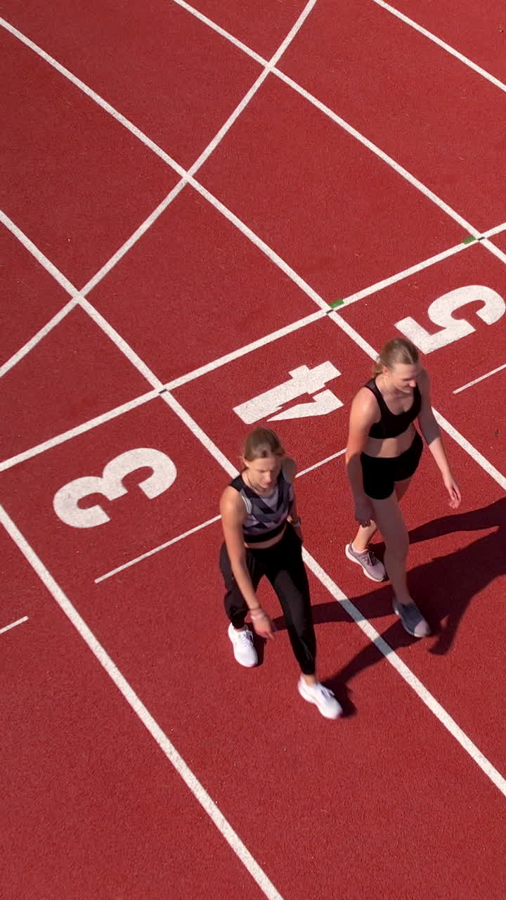 Ripresa dall'alto di due donne atletiche in abbigliamento sportivo che camminano su una pista da corsa rossa con i numeri delle corsie. Ideale per contenuti su fitness, salute e sport