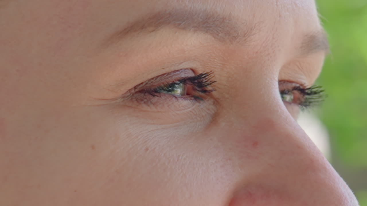 햇빛 아래 공원 배경에서 섬세한 눈매를 가진 사려 깊은 백인 여성의 표정을 담은 클로즈업 이미지를 표현한 것입니다. 야외에서 옆을 응시하는 백인 여성의 모습과 섬세한 눈매가 돋보입니다.