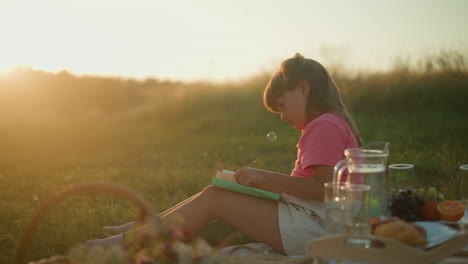 little girl sitting outdoors during sunset peacefully reading book, bathed in warm golden sunlight with bubbles gently dropping on her, surrounding picnic setup with fruits and croissant