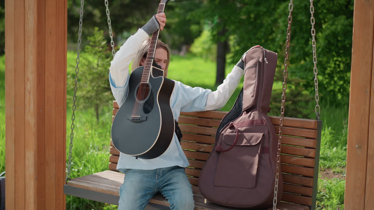 giovane chitarrista bianco seduto su una panchina del parco che apre la custodia imbottita e tira fuori la chitarra acustica, appoggia lo strumento sulle gambe, accorda le corde e suona una dolce melodia, seduto su un'altalena di legno
