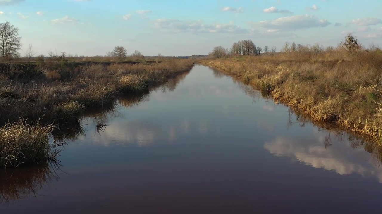 Aerial view moving forward over a placid river with calm, dark water reflecting the blue sky and clouds. The banks are covered in dry, brown grass and sparse trees under a sunny autumn day