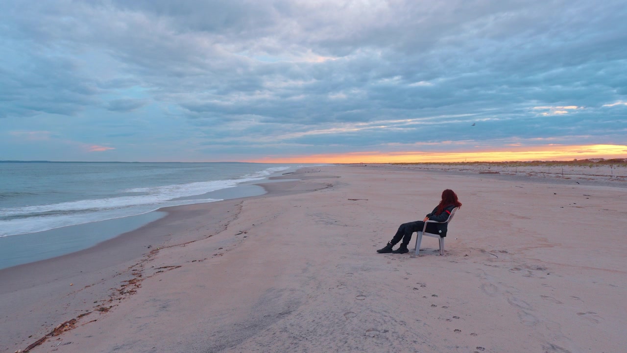 En person sidder i en stol på stranden under solnedgang med bølger og skyer på himlen.