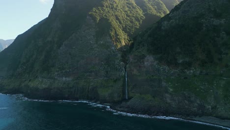 Die-Perspektive-Einer-Drohne-Fängt-Den-Atemberaubenden-Wasserfall-Véu-Da-Noiva-Auf-Madeira-Ein,-Der-Vom-Sonnenlicht-Inmitten-Eines-Bergigen-Geländes-Beleuchtet-Wird-Und-Blick-Auf-Das-Meer-Bietet