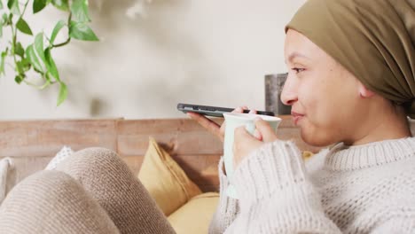 video of happy biracial woman in hijab drinking coffee and talking on smartphone at home