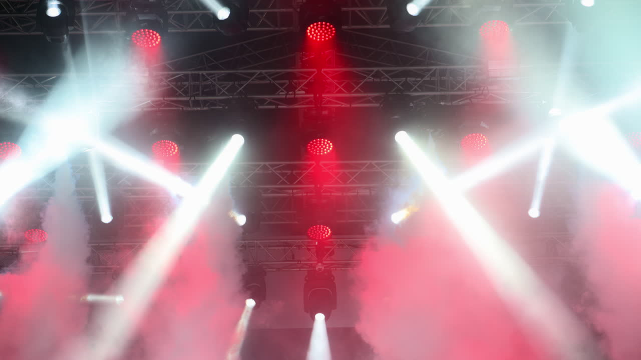 Colorful lights shine down on a lively music event, illuminating the stage with red and white beams while fog adds a dramatic effect to the atmosphere