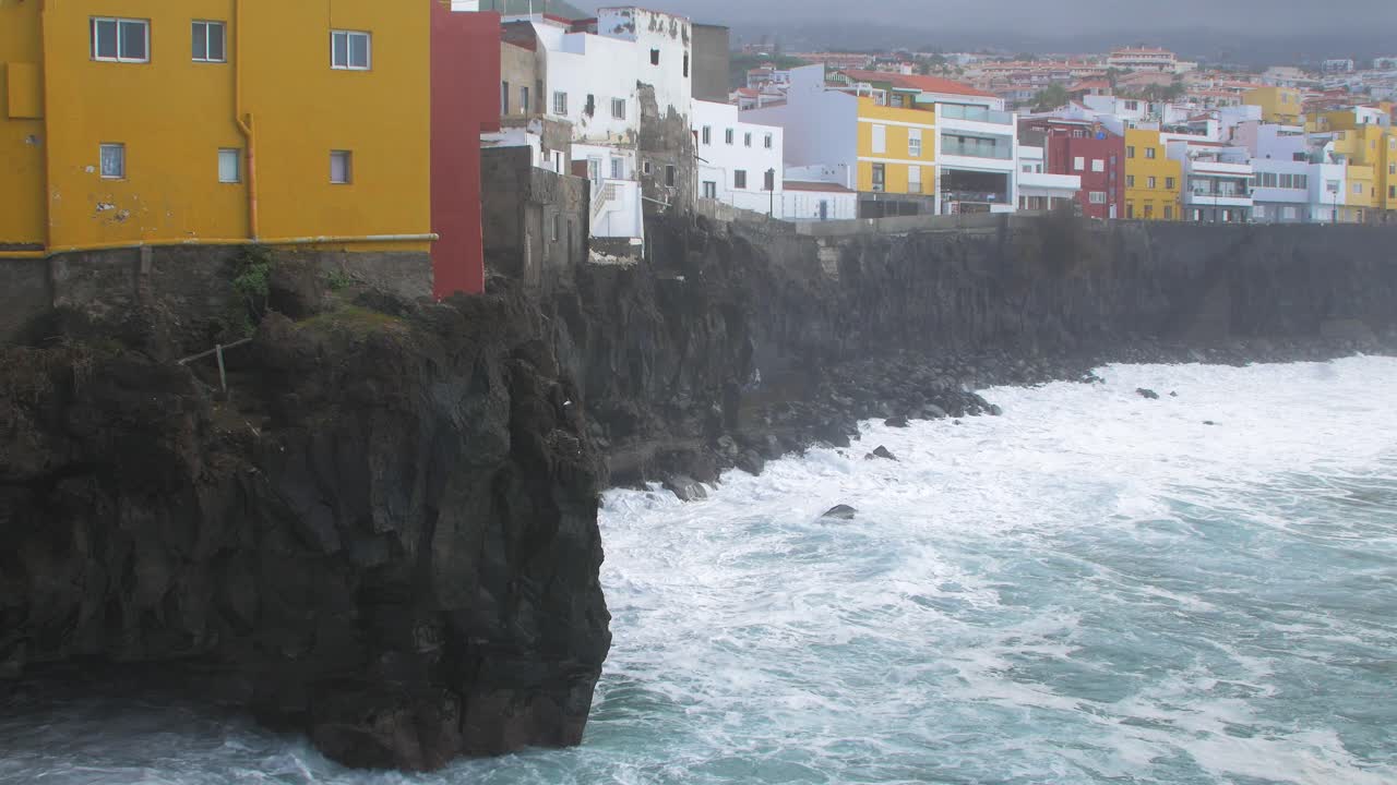 Grandi onde dell'Oceano Atlantico si infrangono su una costa rocciosa in una giornata di sole durante una tempesta a Puerto de la Cruz, Isole Canarie; case colorate in lontananza, ripresa media-ampia.