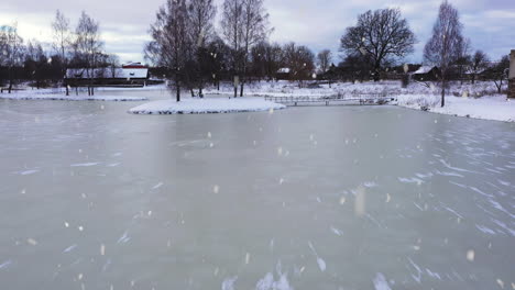 frozen lake in park of small town with snowy landscape while snowing, aerial low altitude view