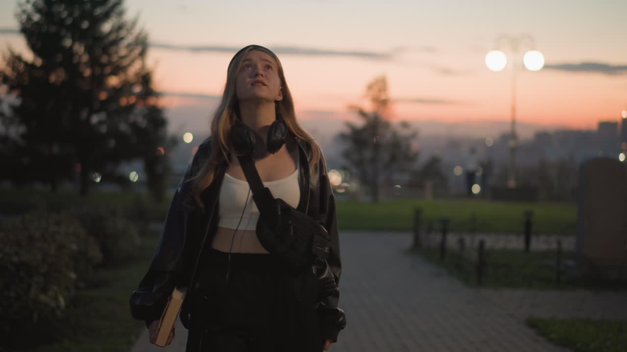 Giovane donna in piedi che guarda la città dall'alto, vista da dietro; si gira e rivela un sorriso mentre il tramonto colora lo skyline, capelli lunghi sotto un berretto, giacca di pelle che fruscia nella brezza, vista panoramica tranquilla, umore contemplativo
