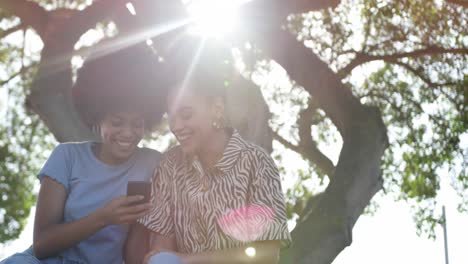 Zwei-Gemischtrassige-Frauen-Schauen-Im-Park-Auf-Ihr-Smartphone