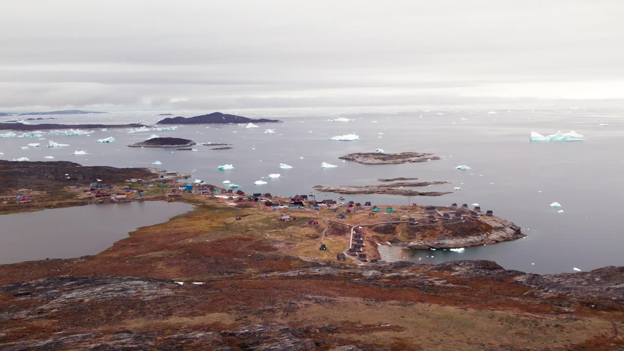 Aerial drone view of the coastal Greenlandic settlement of Ilimanaq, surrounded by Arctic mountains, icy fjords, and the pristine northern seascape