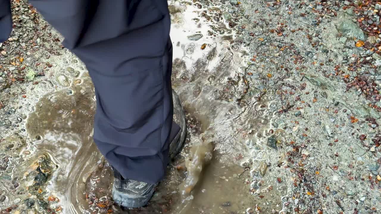 Ein Wanderstiefel spritzt in eine schlammige Regenpfütze auf einem Schotterweg, bei Tageslicht mit Handkamerabewegung und natürlicher Außenbeleuchtung aufgenommen.