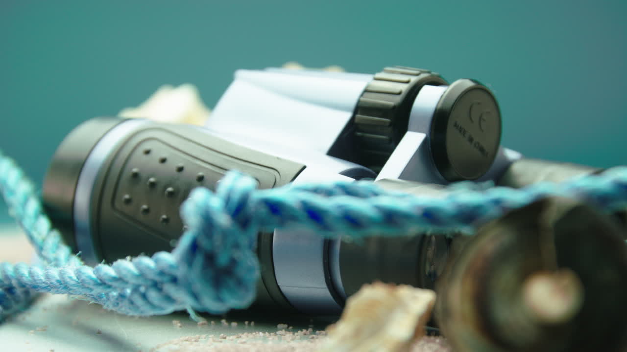 Close-up product video of blue and black binoculars with sea shells and blue rope, surrounded by beach sand, on a turntable display