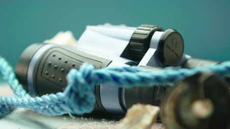 close-up product video of blue and black binoculars with sea shells and blue rope, surrounded by beach sand, on a turntable display