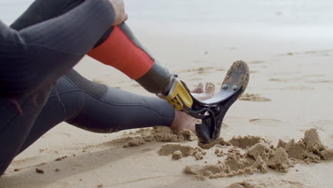 close up of an unrecognizable sportsman with artificial leg sitting on sandy beach and resting after surfing workout in ocean