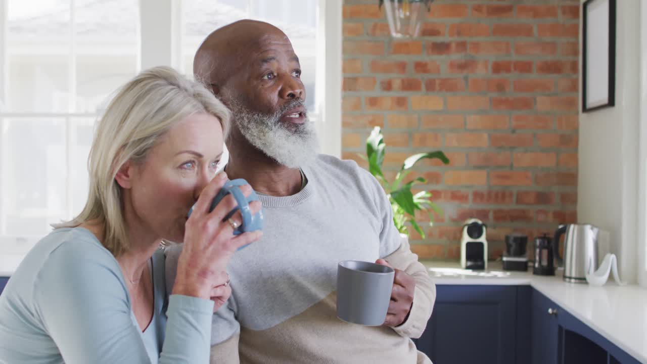 Eldre par med blandet bakgrunn drikker kaffe sammen på kjøkkenet hjemme.
