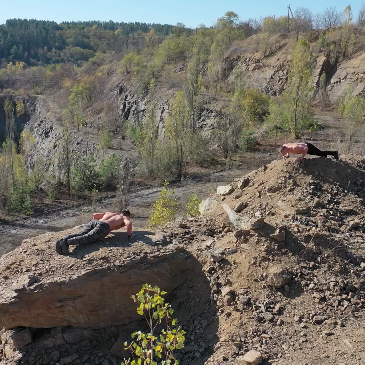 Shirtless sportsmen training on the nature background. Two young athletes doing push ups in the mountainous area in summer day. Aerial view.