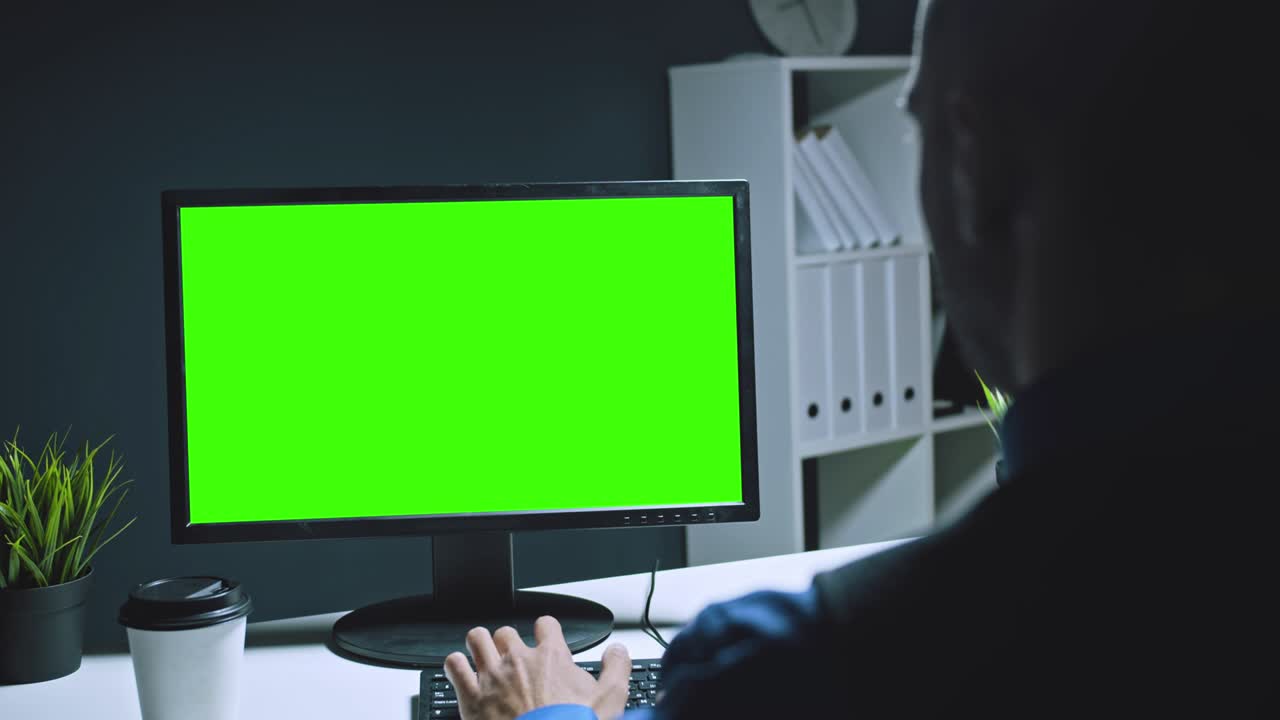 vista trasera de un hombre de negocios exitoso sentado en la oficina y escribiendo en el teclado de la computadora con una pantalla verde vacía