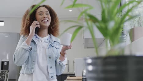 Happy-biracial-woman-talking-on-smartphone,-laughing-and-holding-coffee-in-kitchen,-slow-motion