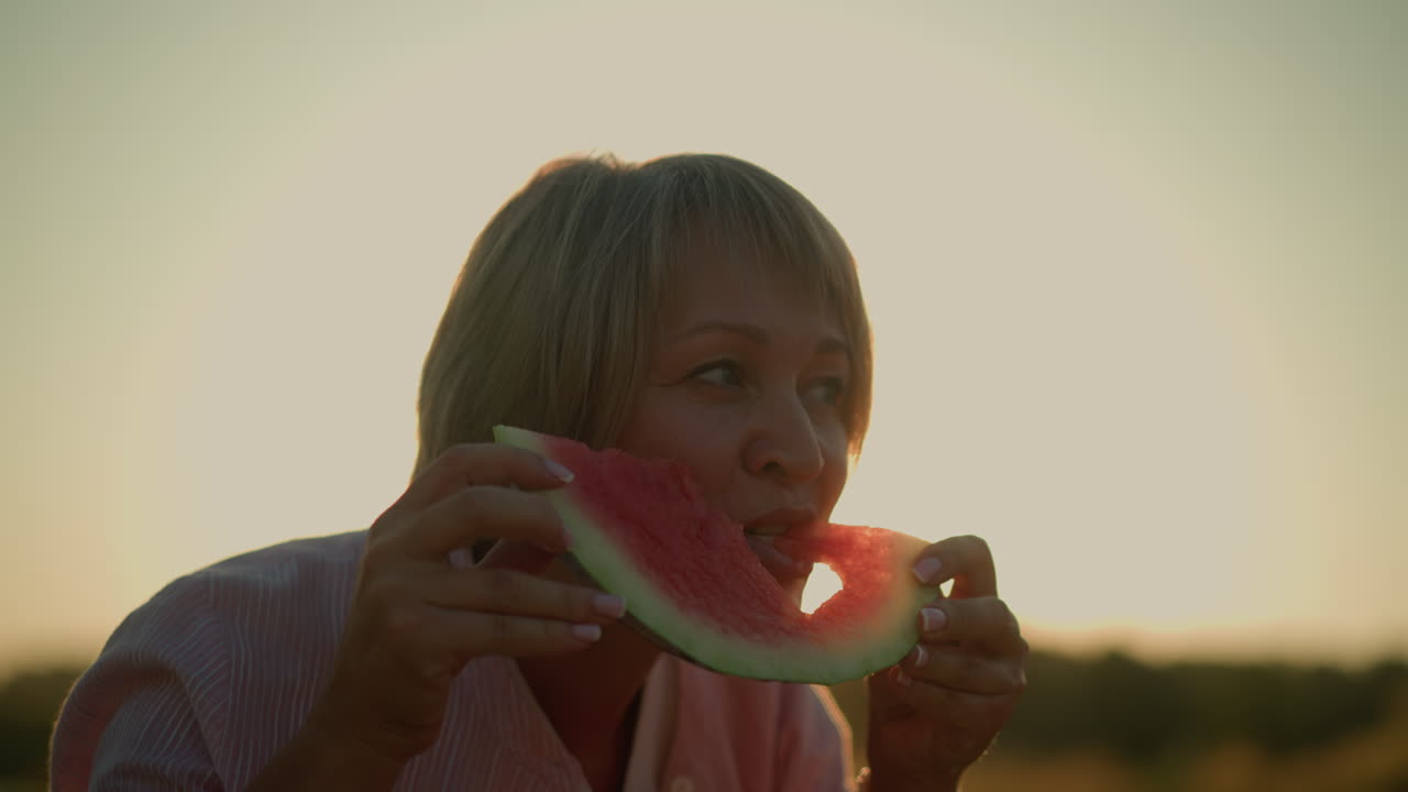 mujer disfrutando de un refrescante bocado de rebanada de sandía con corte en forma de amor bajo un cielo de verano brillante, la luz del sol crea una silueta romántica mientras la fruta roja vibrante