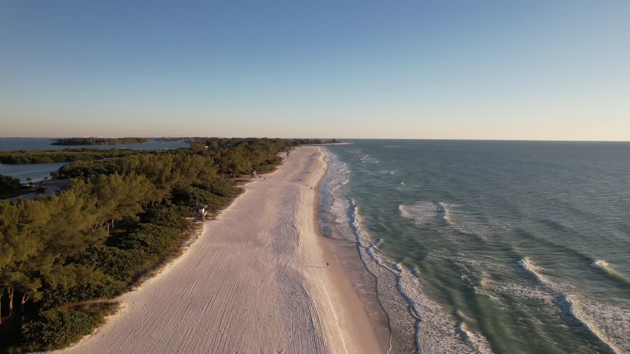 Nagranie z drona poruszające się wzdłuż złocistej piaszczystej plaży z zalesionymi wyspami i falami delikatnie rozbijającymi się o brzeg.