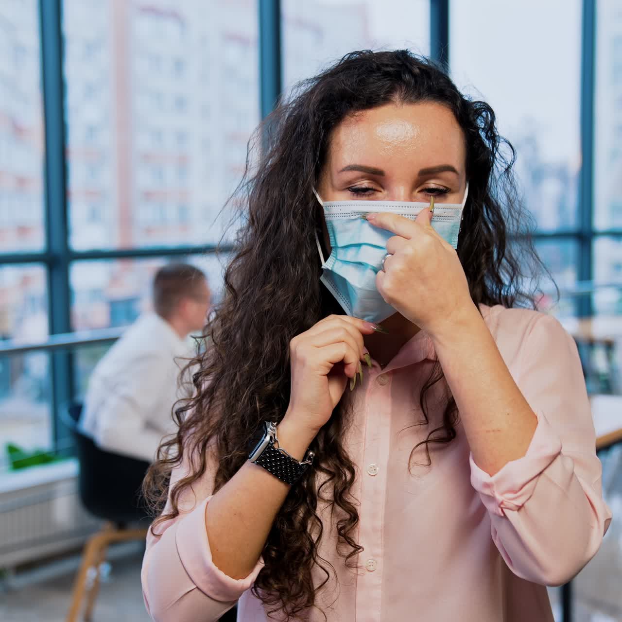 Long-haired lady stand in the office and puts on mask. Office people sitting behind at the table. Panoramic windows at backdrop