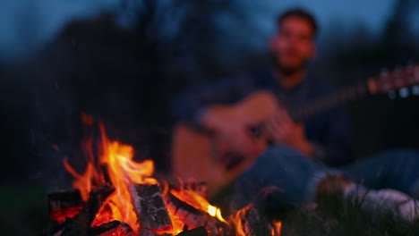 Evening at bonfire. Red and orange fire flame at night. Bright campfire on blur background of a man playing the guitar.