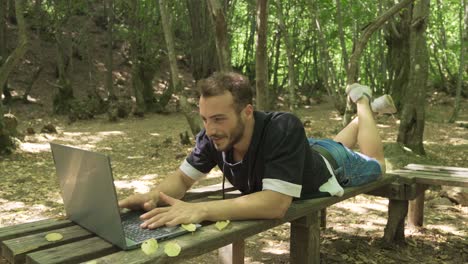 young man lying on a bench in the forest works with a laptop.