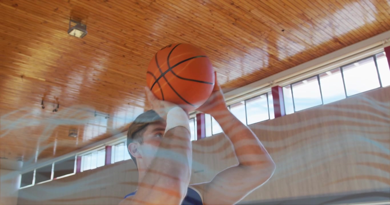 Atleta maschio che finisce di palleggiare un pallone da basket arancione su un campo da palestra, scruta il canestro e lancia un tiro in sospensione.