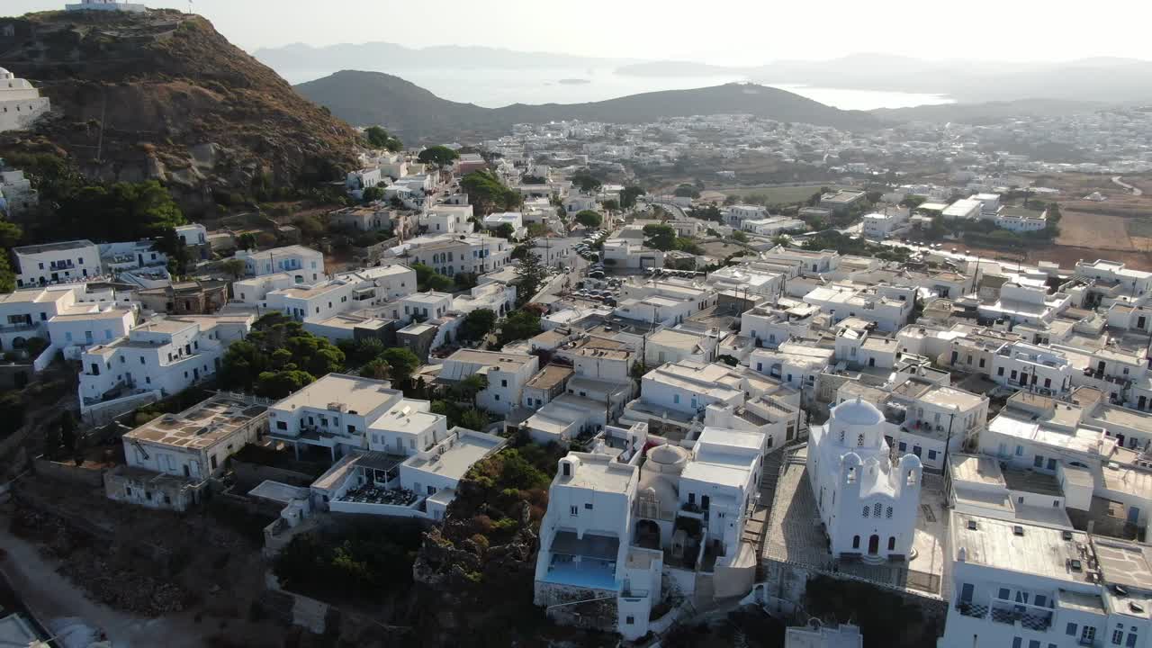 vista de drones en grecia volando frente a la ciudad griega con casas blancas en una colina marrón y el mar en el horizonte en un día soleado