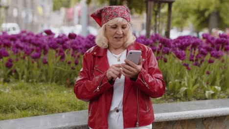 Smiling senior woman using smartphone typing text answering messages chatting online in city street